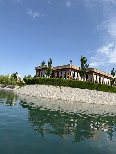 The image, taken in Kuchtumgali, Uzbekistan, depicts a serene outdoor scene under a bright blue sky with scattered white clouds. In the mid-ground, a long, ornate building with traditional architectural elements spans horizontally. It features a patterned roofline, decorative columns, and white fabric drapes or awnings providing shade along its facade, suggesting an outdoor dining area or lounge. Lush green trees are planted around the building, complementing a neatly trimmed green hedge that runs along a low wall or pathway made of small, light-colored pebbles.
To the right, a tranquil body of water, likely a large pool or canal, reflects the building, trees, and sky with a ripple effect distorting the reflections. The water's surface is a deep greenish-blue. No specific activity or people are visible, emphasizing the peaceful ambiance. The time of day appears to be bright daylight, possibly midday or early afternoon, given the clear sky and strong illumination. The architectural style hints at a Central Asian influence, fitting the location. There is no visible text in the image.