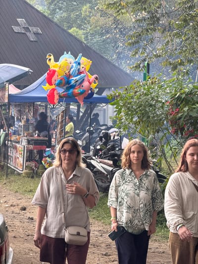 This media file captures an outdoor scene in Banjar, Indonesia, likely a bustling market. Three women are the primary subjects. The woman in the foreground, wearing glasses and a light-colored shirt with a purse slung across her body, looks directly at the camera with a neutral expression. To her left, another woman with blonde hair wears a patterned shirt and holds a dark smartphone. The third woman, partially visible at the top, has reddish-brown hair and is wearing a light-colored shirt.

The scene is filled with various objects: a bunch of colorful balloons shaped like animals and cartoon characters are prominent, some attached to a blue canopy. Behind the women, motorcycles are parked, suggesting transportation for visitors or vendors. Further in the background, market stalls are set up, displaying goods, possibly food items, indicated by shelves filled with what appear to be packaged items and a display case. Smoke or steam rises from somewhere behind the stalls, adding to the market atmosphere. The ground is a mix of dirt and grass.

The lighting suggests it is daytime, with bright sunlight casting shadows. The overall mood appears casual and observational, capturing a snapshot of daily life at a local market. No visible text is clearly discernible in the image.