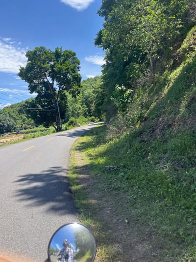 A motorcycle rider is on a paved road in Ham Thuan Bac, Vietnam, on a sunny day with a clear blue sky and scattered clouds. The road curves to the right, bordered by lush green vegetation and trees on the left and a steep, grassy embankment on the right. Shadows of tree branches are cast across the road. In the foreground, a motorcycle's rearview mirror reflects the rider wearing a helmet and a plaid shirt, along with the sky and trees. In the distance, a few more trees and buildings are visible on the left side of the road, and the road continues uphill into a forested area.