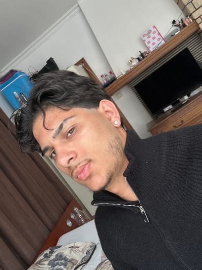 A selfie taken indoors. The subject is a young man with brown hair, brown eyes, and light skin. He wears a black zipped sweater. The background includes a television, wooden furniture, a white wall, and a brown curtain. No evidence indicates this photo was taken in Wetteren, Belgium.
