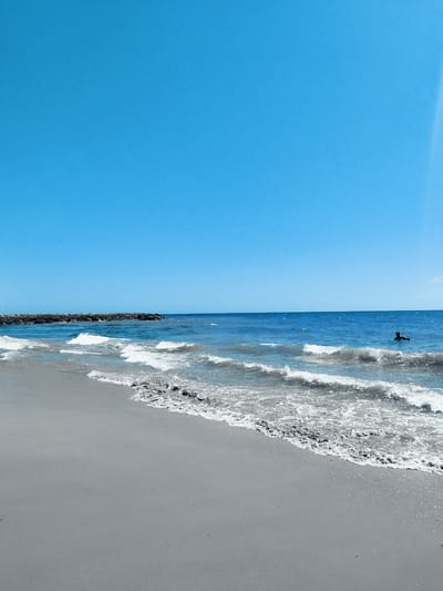 This media file depicts a serene beach scene under a clear, bright blue sky, suggesting a sunny day. The water is a deep blue, with gentle waves lapping onto a gray sandy shore. In the distance, a rock jetty extends into the sea, providing a visual break in the expanse of water. Further out, a lone figure is visible in the water, appearing to be floating or swimming, adding a sense of peaceful solitude to the scene. The overall mood is tranquil and idyllic, evoking a sense of calm and relaxation. The location is identified as Rota, Spain, a coastal town known for its beautiful beaches. There is no visible text in the media file.