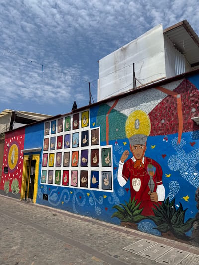 A brightly painted mural on the side of a building in Oaxaca City, Mexico, depicts a figure wearing indigenous dress, and a grid of hand symbols likely representing sign language. The background is predominantly blue with swirls and small birds. Above and to the left, a corrugated metal structure is visible, extending toward a blue sky with scattered clouds. Below the mural, a yellow door and a lower section of mural with an oval design are partially visible. The ground is paved with stone.