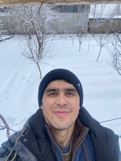 A man wearing a dark blue beanie with a small white logo poses for a selfie outdoors in a snowy landscape. He is bundled in a dark winter coat with visible layers underneath. Behind him, snow-covered ground stretches towards a building and bare trees, suggesting a cold, wintry environment.