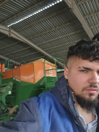 A man with dark, curly hair and a beard is shown in a tilted, close-up shot. He is wearing a blue and grey jacket. Behind him is agricultural machinery, specifically an AMAZONE EDX 9000-TC, painted in green and orange. The background reveals the interior of what appears to be a large shed or barn, with a corrugated metal roof supported by wooden beams. The location is Ovcharovo, Bulgaria.
