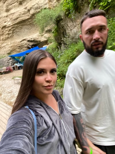 In the city of Salta, Russia, two individuals, a woman and a man, are captured in a close-up selfie. The woman, positioned in the foreground and to the left, has long dark hair and is wearing a grey velour tracksuit with a blue strap visible over her shoulder. The man, to her right and slightly behind, has short dark hair, a beard, and is wearing a white t-shirt. His left arm displays a tattoo. They are standing outdoors with a rocky cliff face and lush green vegetation in the background. Below the cliff, a blue tarp covers what appears to be a resting area with some scattered items. A wooden walkway or platform is visible in the lower left of the frame. The environment suggests a natural, possibly mountainous or canyon-like setting.