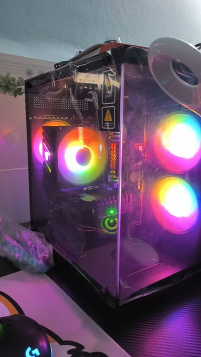A black computer tower with a clear glass side panel is on a desk with a white mouse pad and a black mouse. The computer tower has a variety of colored lights illuminating the internal components. The internal components include a motherboard, CPU cooler, RAM sticks, and graphics card. The tower appears to be on a desk in a home, as there is a houseplant and a tiled wall in the background. The desk appears to be in a home office in Thành phố Hồ Chí Minh, Vietnam.