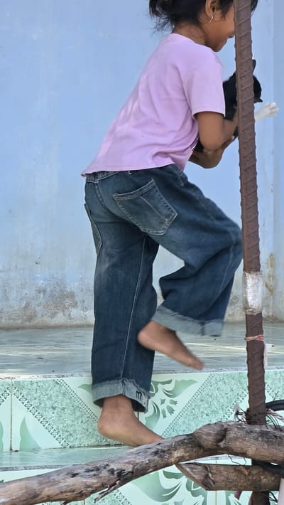 In Ba Ngoi Ward, Vietnam, a child in a pink shirt and blue jeans hangs upside down from a horizontal metal bar. The bar is secured to a weathered wooden structure. The child is also holding onto a black-and-white cat. The background consists of a light blue wall and a tiled floor with a green pattern.