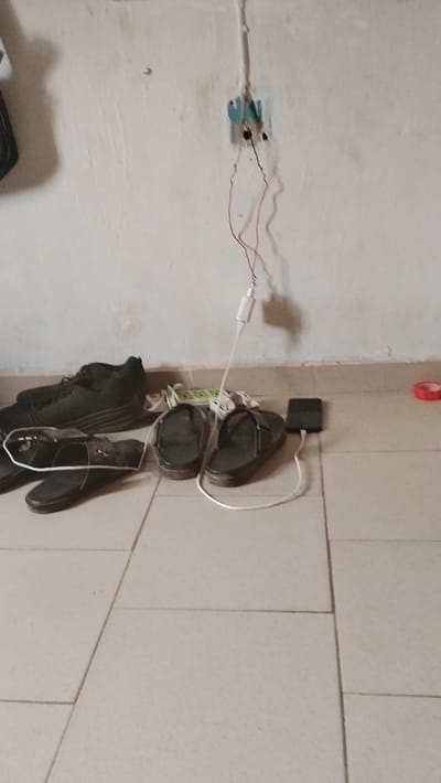 The image depicts an interior scene within a building in Isieke, Nigeria, featuring a light-colored, textured wall above a dark baseboard and a light-tiled floor. An exposed electrical wall socket is visible on the wall, from which two insulated wires, one red and one black, extend. These wires are directly connected to a white USB power adapter. A white USB charging cable extends from this adapter to a dark-colored smartphone lying flat on the tiled floor, indicating the phone is actively charging. To the left of the charging setup, against the wall, are two pairs of black footwear: one pair of athletic shoes and one pair of sandals. Nearby, a green and white object and some crumpled white material are present on the floor. A roll of red electrical tape rests on the floor towards the right edge of the frame. No individuals are visible.