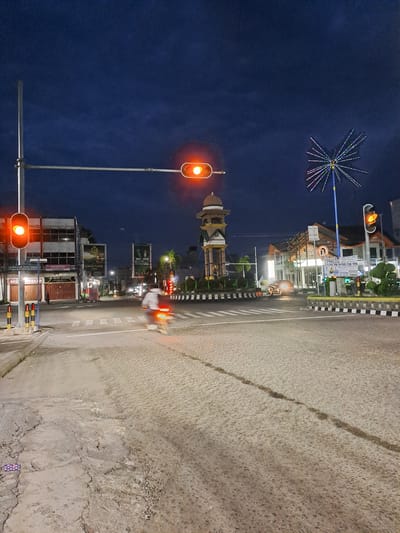 This is a nighttime photograph of an intersection in the city of Bireuen, Indonesia. The sky is dark blue, indicating it is twilight or early night. There are several traffic lights visible, all displaying a red light, suggesting traffic is stopped. In the foreground, the road surface is cracked and appears to be under repair or in poor condition. A motorcycle is blurred in motion, with its taillight illuminated, indicating it is moving through the intersection.
On the left side of the image, there is a traffic light pole with two red lights. Behind it, there are buildings, including one with a sign that reads "smartfren." Further down the street, there are more buildings and streetlights.
In the center of the image, a distinctive monument or clock tower stands, topped with a dome and adorned with ornamental details. It is illuminated by lights. To the right of the monument, there is another traffic light, also red. Beyond that, there is a street with buildings and a unique, star-shaped decoration with colorful lights. A car is visible in the distance, its headlights on.
The overall scene depicts a busy urban intersection at night, with elements of infrastructure, traffic control, and a local landmark. The condition of the road suggests ongoing construction or maintenance.