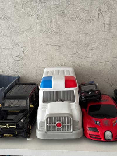 A collection of toy vehicles is displayed on a white shelf against a textured, light-colored wall. From left to right, the first visible toy is a black SUV, resembling a Mercedes-Benz G-Class, with gold accents and a license plate that reads "M923Y". To its right is a large, white toy truck with blue and red lights on its roof. Stacked on top of the white truck is a black toy police car, a Ford Mustang, with blue and red emergency lights. Next to the white truck and partially obscured by it is a red toy sports car, resembling a Bugatti, with various decals and markings. The background wall has a repeating pattern of raised, irregular lines. The lighting appears to be from above, casting subtle shadows on the shelf and wall. There are no people or discernible signs of human activity in the immediate scene. The location is specified as Yarominski Sel'ski Sovet, Belarus.