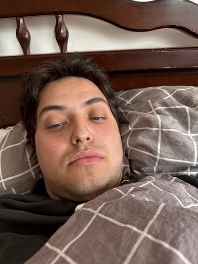 A man is lying in bed under a gray and white plaid blanket. He has dark hair, light skin, and blue eyes. He appears to be looking directly at the camera. The headboard of the bed is visible to the left and is made of dark wood. The location is Гаспринский поселковый совет, Ukraine.
