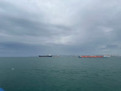 The image depicts a seascape under a cloudy sky in Unknown, Türkiye. Several ships are visible on the water. In the distance, a coastline with buildings is discernible. The sea is relatively calm, with small waves.