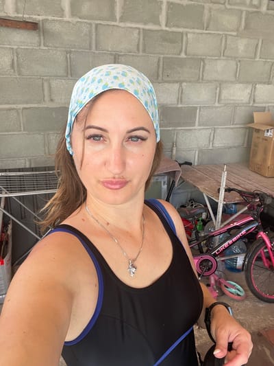 A woman is taking a selfie outdoors, with a brick wall to her left and a cluttered area with bicycles and storage to her right. She is wearing a black athletic top with blue trim and a blue and white patterned bandana on her head. She has on a silver necklace with a charm. Her expression is neutral, and the lighting suggests it is daytime. The background includes a pink bicycle, a smaller pink scooter, and some cardboard boxes. The scene appears to be a backyard or outdoor storage area. No visible text is present in the image.