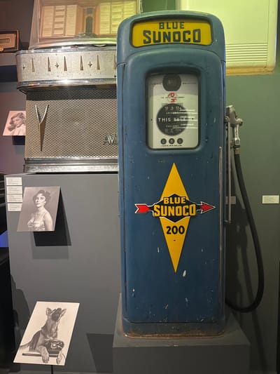 The image shows a vintage blue Sunoco gas pump with a yellow "Blue Sunoco" logo and the number "200". The gas pump is located in a museum display. To the left of the gas pump are several framed photographs and what appears to be a vintage jukebox. The photographs show a woman in a white top, another woman in a formal dress, and a German Shepherd dog with its paws on a rotary telephone. The background is a dark gray wall. The gas pump is standing on a dark gray platform. The image does not provide any information about the city of Bern, Switzerland.