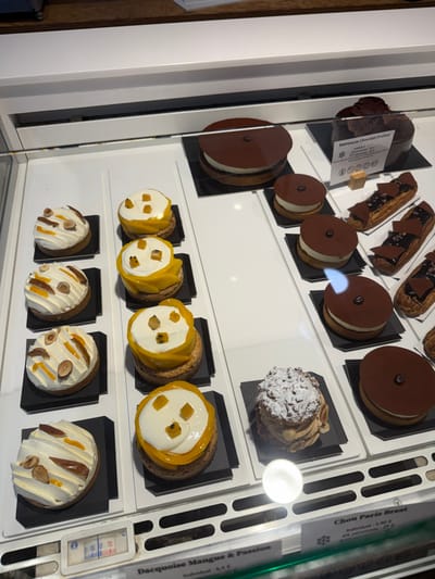 A display case presents rows of assorted pastries. The top row features chocolate-glazed confections and eclairs. The middle rows showcase round pastries with white cream centers and colorful fruit toppings. The bottom row has similar round pastries adorned with cream, nuts, and orange segments. Each pastry rests on a small, square, black platform, neatly arranged within the illuminated, white interior of the display case.