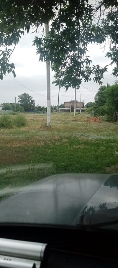 The image shows a view from inside a vehicle looking out onto a grassy area with some trees and buildings in the background. The foreground is dominated by the hood of the vehicle, which is dark and reflective. In the middle ground, there is a patch of dry grass and a pile of dirt. In the background, there are several houses and a larger building with a distinctive roof. Two poles with wires are visible in the middle ground, suggesting a rural or semi-rural setting. The sky is overcast. The scene appears to be in a residential area, possibly on the outskirts of a town or village. The location is specified as Marinovka, Kazakhstan.