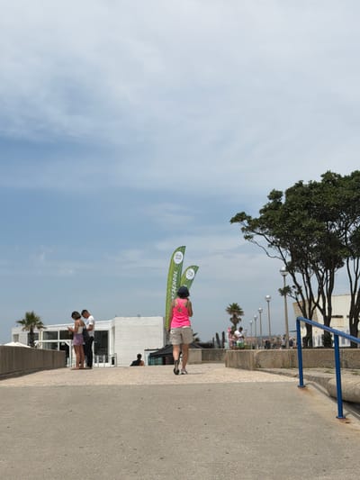 In Espinho, Portugal, on a paved walkway, a woman with short dark hair, wearing a bright pink tank top and light-colored shorts, is walking away from the camera towards the right. Her back is to the viewer, and she is wearing athletic shoes. In the middle ground, on the left, a man and a woman are standing close together, both looking down at their phones. The man is wearing a white t-shirt and dark pants, and the woman is wearing a white tank top and light-colored shorts. Behind them, a modern, white building with large windows is visible. To the left of the building, a palm tree stands tall. In the background, several green, vertical banners are displayed, one of which reads "SURF SCHOOL". Further in the background, trees and other buildings are present under a cloudy blue sky. A blue railing runs along the right side of the walkway.