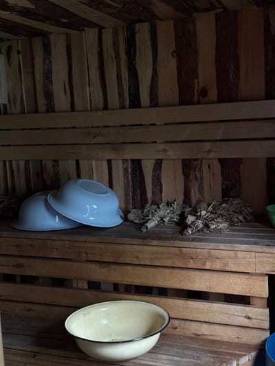 The image shows a rustic wooden interior, likely a sauna or banya, located in Zykova, Russia. The walls and ceiling are constructed from unvarnished wooden planks of varying widths and natural bark edges, creating a textured and organic aesthetic. Two levels of wooden benches are visible. On the upper bench, two light blue enamel bowls are stacked, with the top bowl inverted over the bottom one. Next to the bowls, on the same bench, are several bundles of dried leafy branches, resembling bath whisks (veniki), some tied with white string. On the lower bench, a single, larger light yellow enamel bowl with a dark rim is positioned centrally. The overall scene evokes a traditional and natural atmosphere common in Russian bathhouses.