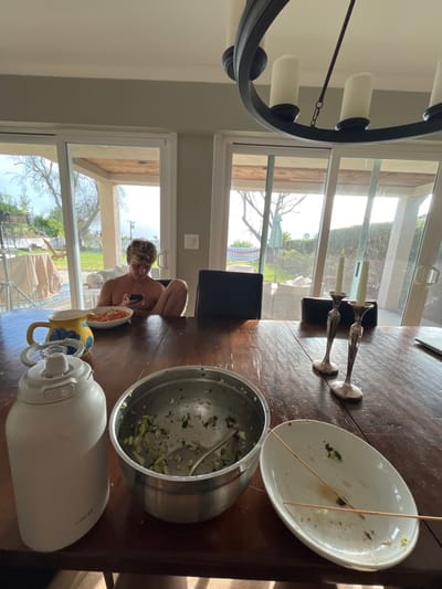 The image displays an indoor dining area within a residence in Rancho Palos Verdes, United States. A large, dark-stained wooden table occupies the foreground and mid-ground. At the far end of the table, a young male, shirtless with light-colored, curly hair, sits using a smartphone held in both hands. His knees are bent and elevated towards the table.
On the table, from left to right in the foreground, are a white insulated jug, a stainless steel mixing bowl containing remnants of chopped green vegetables and a spoon, and an oval white serving platter with food residue and two wooden skewers. Further along the table, to the left, is a yellow ceramic pitcher with a blue floral design, and a smaller white, lidded container. Closer to the seated individual, a white bowl holds orange-colored food. To the right, two silver candelabras each hold an unlit white taper candle. Behind the candelabras, a dark-colored laptop or tablet with a closed screen is positioned, alongside another dark electronic device.
The room has light-colored walls and features a dark, ornate metal chandelier with white, candle-shaped light covers suspended from the ceiling. Large glass sliding doors and windows dominate the background, revealing an outdoor patio with furniture (including a towel-draped object) and a grassy yard beyond. The yard contains trees, shrubs, a distant white fence, and a blue object. The scene is illuminated by natural light from a bright sky.
