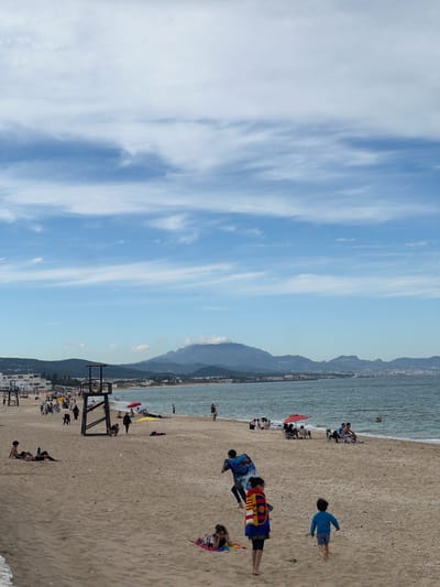 The image captures a coastal scene at M'Diq, Morocco, showcasing a beach populated with people. On the sandy beach, several individuals are visible, including some lying down, others standing, and one running. Adjacent to the beach is the Mediterranean Sea, with a coastline stretching into the distance. In the background, mountainous terrain adds depth to the scene. The sky above is partially covered with clouds. Beach umbrellas and what appear to be lifeguard structures are also visible along the shoreline.