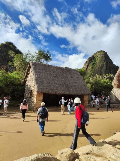 Tourists explore an open archeological site on a sunny day with a partly cloudy sky. A central stone structure with a thatched roof stands prominently. Several people are either walking around the site or standing near the building, some wearing hats and backpacks. Lush green hills and trees surround the open area.