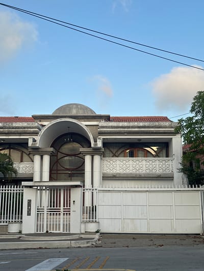 The image shows the facade of a large, ornate house with a dome, located at 42-60 on a street in Perímetro Urbano Barranquilla, Colombia. The house is painted white and features arched windows with decorative metalwork, columns, and a tiled roof. A white metal fence and gate system encloses the property. Power lines are visible across the sky, which is partly cloudy. The street in front of the house is paved, with a crosswalk and speed bumps. There are no people visible in the image.
