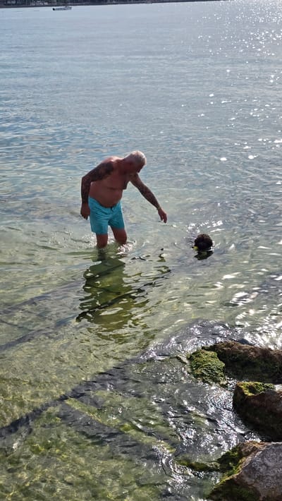 A man with tattoos on his arms and light-colored hair is standing in clear, shallow water near a rocky, moss-covered shoreline. He is wearing light blue swim trunks. His left arm is extended forward as if reaching out. The water is shimmering with sunlight, and the bottom is visible, showing a rocky and green-algae covered surface. In the background, the water stretches out with light reflections. A dark, round object, possibly a diving mask or a hat, is floating in the water to the man's right. The scene appears to be outdoors on a sunny day, likely in the afternoon given the angle of the light. The location is identified as Peschiera del Garda, Italy. There is no visible text in the image.