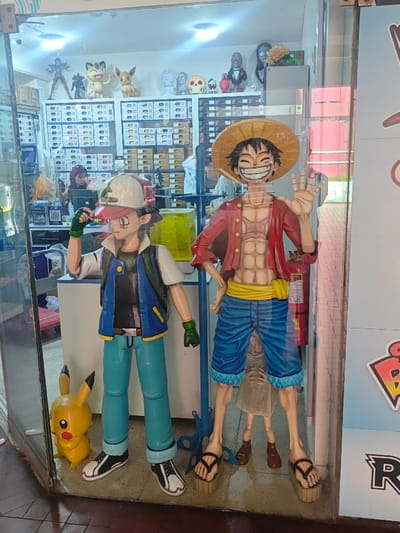 The image displays the brightly lit storefront of an anime and collectible shop, likely located in Lince, Peru. Dominating the display are two large, life-sized or near life-sized statues of popular anime characters behind a glass panel. On the left stands Ash Ketchum from Pokémon, identifiable by his white and red cap, blue vest, and green-gloved hands, one of which adjusts his cap. To his right is Monkey D. Luffy from One Piece, sporting his iconic straw hat, red open shirt revealing his chest, blue shorts, and sandals. He is depicted with a wide, cheerful grin and a raised hand, as if waving. A small yellow Pikachu figurine is positioned on the floor next to Ash.
Through the glass, the interior of the store is visible, featuring multiple shelves stocked with various merchandise boxes. Above these shelves, several smaller character figures and busts are displayed, including Meowth and Eevee from Pokémon, along with what appear to be figures from "The Nightmare Before Christmas" and decorative skulls. Two individuals can be seen inside the shop; one person is seated at a counter on the left, while another is partially visible further back on the right, indicating the store is open.
Notable details include significant reflections on the glass, such as ceiling lights and a distorted reflection of a small child's lower body appearing between Ash and Luffy. A yellow container is visible behind Ash, and a red fire extinguisher stands behind Luffy. On the far right of the display, partial, stylized text is visible, including a prominent 'R' and another letter resembling a 'P' or 'B' with a pinkish-red outline, likely part of the store's branding or logo.