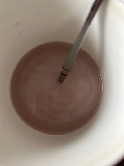 A top-down, close-up shot shows a white mug filled with a light brown liquid, possibly hot chocolate or coffee with milk. A silver spoon is partially submerged in the liquid, with a visible swirl pattern on the surface of the drink, suggesting it has been recently stirred. The mug appears to be on a flat surface, and the lighting is soft and diffused. The background outside the mug is out of focus and appears to be a lighter color, possibly a table or countertop. The image does not provide any specific details about the city of Kaa-Khem, Russia, or any individuals present.