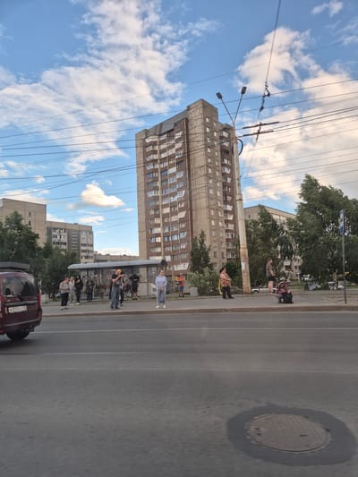 The image depicts a street scene in Petrozavodsk, Russia, with a focus on a bus stop and an apartment building. A tall, weathered apartment building with numerous windows dominates the background, slightly tilted. To its left, another, shorter apartment building is visible. In the foreground, a road with a manhole cover is present, and a maroon van with a license plate reading "M395CO 10" is parked on the side.

At the bus stop, several individuals are gathered. Some are standing, while others are seated under the shelter. One person is holding a camera and appears to be taking a photo. Another person is kneeling. The people are dressed in casual attire. Overhead, power lines and streetlights crisscross the sky, which is partially filled with scattered clouds against a blue backdrop. The overall impression is of a typical urban environment.