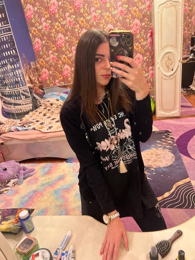 A woman with long brown hair and dark clothing takes a selfie in a bedroom. She is wearing a black long-sleeved shirt with a graphic print and multiple necklaces. A watch with a white and gold band is visible on her left wrist. The background of the room includes a bed with patterned bedding, a floral wallpaper, and a tall, ornate wardrobe. A colorful, fluffy rug is on the floor in front of the bed, with a purple octopus plush toy on it. On a surface in the foreground, there are various toiletries, including tubes of toothpaste and a hairbrush. The image appears to be taken in a residential setting.