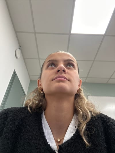The image displays a head-and-shoulders portrait of a female subject, observed from a low-angle perspective, situated indoors in what appears to be a modern institutional or office environment in Saint-Étienne, France. The subject, a young woman, looks upward with a neutral expression. She has light-colored eyes, prominent eyelashes, and light brown or blonde curly hair visible around her ears and shoulders. She is wearing a black textured cardigan over a white collared shirt with eyelet details. Gold-toned floral or clustered earrings are present on both ears, and a gold-toned necklace with a small, flower-shaped pendant rests at her collarbone. The background consists of a white wall on the left, where a white circular device, possibly a sensor or detector, is mounted near a thin dark cable. A light green door frame or panel is partially visible on the far left. The ceiling is a white suspended grid system with several rectangular, illuminated LED light panels casting bright white light.