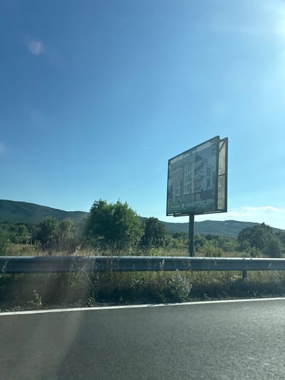 The image displays a billboard advertising real estate, likely an apartment building, against a backdrop of a clear blue sky and rolling green hills. The billboard features an architectural rendering of a modern building with multiple stories and balconies. Text on the billboard includes "omalaa," what appears to be a brand or project name, and a phone number "+359 877 507 517". There is also text in a foreign language, possibly Cyrillic, and a price listed as "€53.000".
The scene is a roadside setting, with a highway barrier and a paved road visible to the right of the billboard. The surrounding environment is lush with green trees and vegetation, suggesting a rural or semi-rural location. The sun is bright, indicating it is daytime, likely late morning or afternoon. The clear sky and warm lighting suggest fair weather. The image is oriented vertically, with the billboard appearing slightly tilted. There are no people visible in the image. The overall impression is one of an advertisement placed strategically along a travel route, likely to attract potential buyers for the advertised property. The location context provided is Dyuni, Bulgaria.
