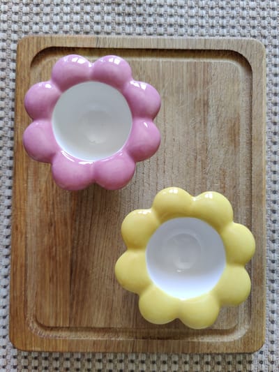 Two flower-shaped ceramic dishes, one pink and one yellow, are placed on a wooden cutting board. The cutting board has a shallow groove around its edge and is situated on a textured, light-colored rug. The scene is captured from a top-down perspective. There are no people visible in the image, and no specific location in Rybnitsa, Moldova, can be identified from the image itself.