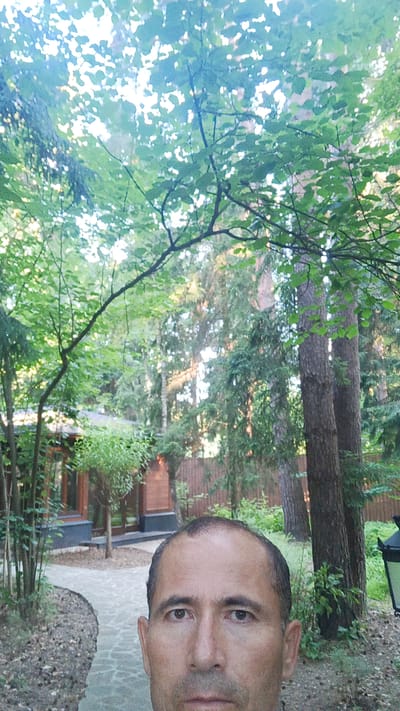 This is a selfie taken by a man in an outdoor setting, likely a garden or park. The man is positioned in the foreground, with his face filling a significant portion of the lower half of the frame. He has short dark hair, brown eyes, and is looking directly at the camera with a neutral expression.
Behind the man, the scene is dominated by lush greenery. Tall trees with dense foliage are visible, with sunlight filtering through the leaves, suggesting it is daytime. In the middle ground, a modern building with large windows and dark-colored trim is partially visible, set within a landscaped area. A winding path made of light-colored paving stones leads towards the building.
Further in the background, more trees and a wooden fence can be seen. The overall atmosphere is serene and natural, with a hint of residential architecture. The time of day appears to be late afternoon or early evening, given the quality of the light. There is no visible text in the image.