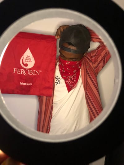 This image, captured in Gbawe, Ghana, depicts a male individual framed by a black ring light. The subject wears a reversed black mesh baseball cap, a red bandana with white paisley patterns covering the lower face from the nose down, a white t-shirt featuring a black atomic symbol graphic, and an unbuttoned red-and-white striped long-sleeved shirt. His right hand holds up a red fabric bag, partially obscuring the left side of his body and face. The bag displays a white blood drop logo containing the text 'THE IDEAL BLOOD BUILDER,' along with 'FEROBIN®' and 'fidson.com.' His left arm is bent, with the hand positioned near the back of his head/neck. The background consists of a plain, light-colored wall. A finger is visible at the lower-left edge of the ring light's frame.