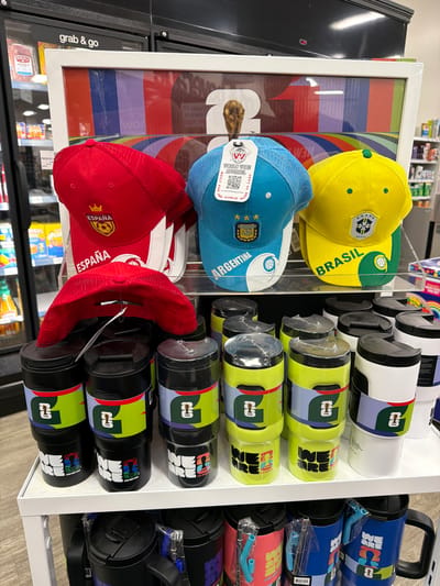 The image shows a display of merchandise, likely in a convenience store or a sporting goods store, in Glendale, United States of America. The merchandise includes baseball caps and tumblers featuring national flags and team logos, with a focus on soccer. Three baseball caps are prominently displayed at the top, one red with "España" (Spain) and a soccer crest, one blue with "Argentina" and the Argentinian national soccer team logo, and one yellow with "Brasil" and the Brazilian national soccer team logo. Below the caps, shelves are filled with cylindrical tumblers. The tumblers are primarily black and white, with colorful bands featuring the FIFA World Cup logo and various slogans like "WE ARE" and city names, suggesting they are related to the World Cup. The background shows refrigerated display cases with food items and shelves stocked with more products, consistent with a retail environment. There are no people visible in the image.