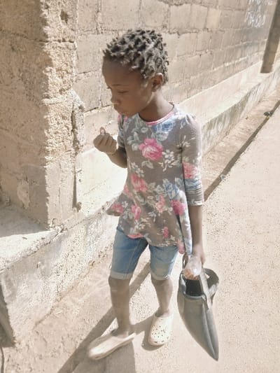 A young child with dark skin and braided hair is depicted walking on a dusty path in Masvingo, Zimbabwe. The child wears a grey long-sleeved floral top with pink and blue patterns, light blue denim shorts, and mismatched clogs (a plain light-colored clog on the left foot, a white perforated clog on the right foot). The child's right hand holds a small, light-colored object near the mouth, while the left hand carries a dark, conical container with a handle, inside of which a clear plastic bottle is visible. The child's gaze is directed downwards. To the left of the child is a rough concrete pillar. Behind the child and extending to the right, a light-colored concrete block wall runs horizontally, featuring a narrow concrete drainage channel at its base. The outdoor scene is brightly illuminated by natural sunlight.