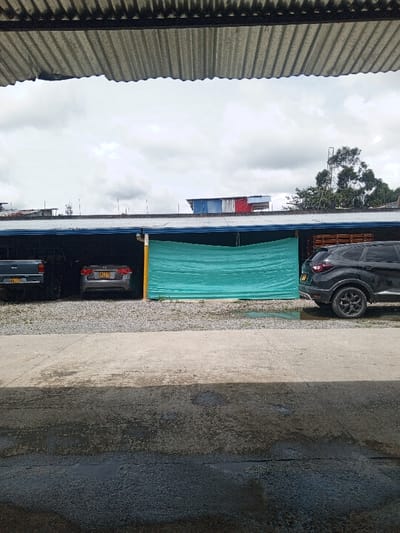 The image presents an outdoor scene viewed from underneath a corrugated metal roof, looking towards a semi-covered parking or storage area under an overcast sky.
In the immediate foreground, a dark, unevenly wet concrete or asphalt surface spans the width of the frame, showing varied tones of grey and black due to moisture. This wet area is separated by a clear horizontal line from a lighter, drier strip of pavement that runs across the mid-foreground.
Beyond this pavement, the ground transitions to a light-colored gravel surface, where a long, low-slung, dark-colored structure provides shelter. This structure has a flat, dark roof, casting a deep shadow over the parking area beneath it.
Underneath the covered area, several vehicles are parked:
On the far left, the rear of a dark-colored pickup truck is partially visible. Next to it, a silver or light grey sedan is parked, facing away from the viewer. Its rear license plate, "LLQ 632," is clearly legible. To the right of the sedan, a bright turquoise or teal-colored tarp or mesh screen is stretched horizontally, acting as a partition or temporary wall, obscuring the area behind it. This screen is supported by a visible yellow pole on its left side. On the far right, the rear and left side of a dark grey or black SUV, equipped with black multi-spoke wheels, is parked, facing towards the right side of the frame. A yellowish-white drainpipe or conduit is visible running vertically down the structure near the left edge of the turquoise screen.
Above the roofline of the parking structure, a vast, light grey, overcast sky dominates the upper background, indicating a cloudy daytime setting. Against this sky, several distant elements are visible:
On the left, faint outlines of what appear to be antennas or communication towers, along with parts of other building roofs, can be seen. In the center-right, a distinct corrugated metal roof with sections painted in blue, white, and red is visible, suggesting a multi-colored building. Further to the right, a tall, sparse tree and another metal structure, possibly an antenna, rise above the distant rooflines.
The very top of the image is framed by the underside of a corrugated metal roof, showing its ribbed texture and signs of weathering, suggesting the photograph was taken from within a similar sheltered area. The diffuse lighting and wet ground indicate an overcast day, possibly with recent rain.