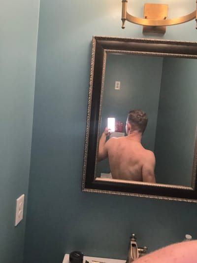 The image shows a man taking a mirror selfie in a bathroom. He is facing away from the camera, showing his back. He is holding a smartphone in his right hand, which is reflecting light. The bathroom has teal walls and a dark framed mirror above a sink. A gold light fixture is visible above the mirror. A white electrical outlet is on the left wall. The image appears to have been taken in Panama City Beach, United States of America.