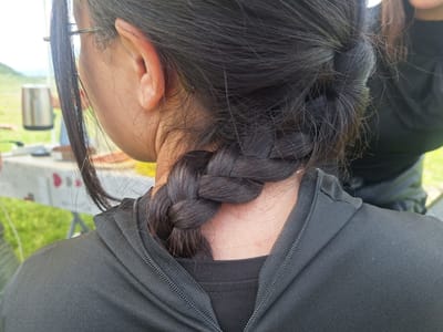 The image shows the back of a person with dark hair styled in a braid that starts high on the head and goes down to the nape of the neck. The person is wearing a black jacket with a zipper visible on the collar. Part of their ear and glasses are visible on the left side. The background is blurry but appears to be outdoors, with green grass and a table visible. There's no indication of a location in the city of Tlik, Armenia.