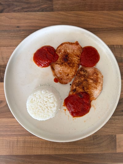 A top-down shot captures a prepared meal served on a round, off-white ceramic plate with subtle dark speckles. The plate rests on a warm-toned wooden surface, displaying a distinct grain pattern. The main components of the meal include two pieces of pan-fried or grilled meat, likely pork chops, characterized by their browned and slightly seared surfaces. These are accompanied by three dollops of a rich, vibrant red sauce, possibly tomato-based or a similar condiment, artfully placed both on and around the meat. Completing the dish is a neatly molded mound of fluffy white rice, positioned towards the bottom right of the plate. The overall presentation is simple and unadorned, suggesting a ready-to-eat, home-cooked or casual dining experience. No people or text are visible in the image. The setting implies an indoor environment, such as a kitchen or dining area.