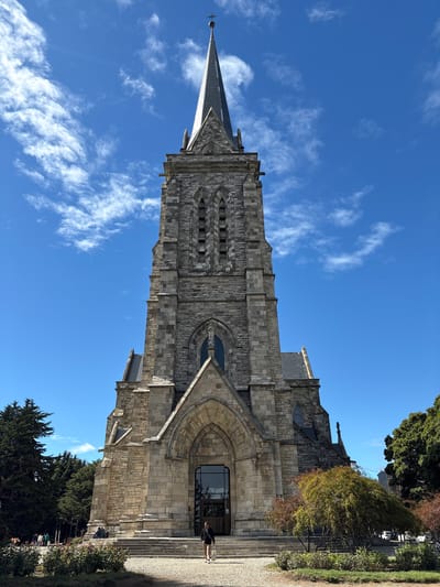 The image features the Cathedral of Our Lady of Nahuel Huapi in San Carlos de Bariloche, Argentina. The cathedral, built in a neo-Gothic style from grey stone, fills the majority of the frame. The structure comprises a tall bell tower with a pointed spire at the top. The cathedral has multiple levels and stone detailing.

A bright blue sky streaked with light white clouds appears behind the cathedral. To the side of the cathedral are trees and greenery. Two people are visible on the steps of the cathedral's entrance.