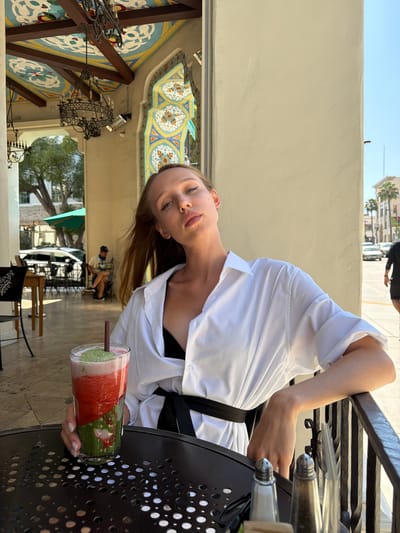 A young Caucasian woman with long, light brown hair and blue eyes is seated at an outdoor table in Pasadena, California. She is wearing a white oversized button-down shirt with the sleeves rolled up and a black belt cinched at her waist. A black bra is visible underneath the shirt. She is leaning on a black metal railing, with her left arm resting on it. Her gaze is directed upwards and slightly to her right.

In front of her on the table is a tall glass filled with a layered red and green smoothie, with a straw in it. The table is round and black with a textured surface. Two salt and pepper shakers are visible on the table.

The background shows an outdoor seating area with tables and chairs. In the middle ground, another person is seated at a table, partially obscured by trees. Beyond that, there are buildings and palm trees, suggesting an urban environment. The architecture features ornate tile work and wrought iron elements. The sky is clear and blue. The scene appears to be a sunny day.