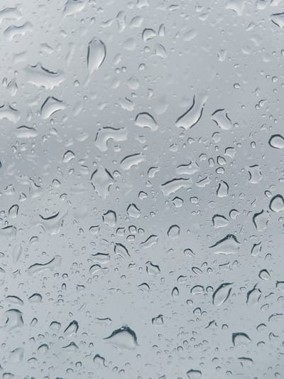 This is a close-up, abstract shot of raindrops on a window. The water droplets vary in size and shape, some are large and elongated, others small and circular. The background is a soft, blurred gray, suggesting an overcast sky or a dimly lit indoor environment. There are no people or specific objects visible that would indicate a particular location within the city of Orlando, United States of America. The image focuses solely on the natural phenomenon of water on a surface.