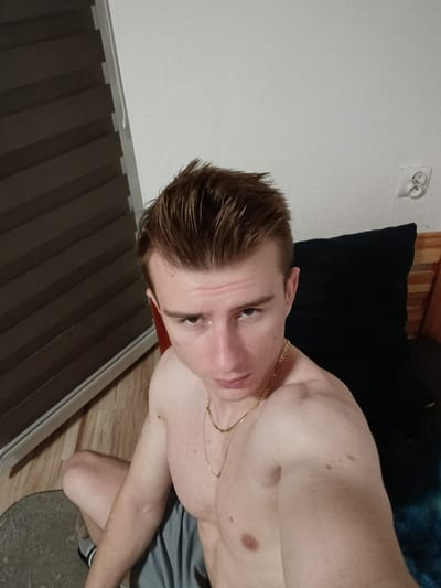 The image displays a young, shirtless Caucasian male with light skin and light brown hair, sitting on the floor indoors. He is wearing a gold chain around his neck and grey shorts. The background features a wall with a white electrical outlet, dark blinds on the left, and a dark pillow behind the subject. The floor is made of wood with a rug in the foreground. The lighting suggests an indoor setting, possibly a bedroom. The image is a selfie taken from a high angle. No specific location in Sucha Beskidzka, Poland is identifiable from the visual evidence.