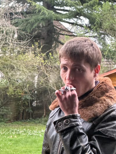 This image depicts a male individual outdoors in Le Raincy, France. The subject has short, brown hair styled with a fringe and light-colored eyes, directed towards the viewer. He is wearing a dark, possibly leather, jacket with a prominent brown fur collar and a dark garment underneath. A single small hoop earring is visible in his right ear. His right hand is raised, with fingers near his mouth. This hand is adorned with multiple rings, including one with a large dark stone on the ring finger, a spiral-designed ring on the middle finger, and a lighter-colored ring on the pinky finger. The background consists of an outdoor environment, likely a garden or backyard. Green grass with small white flowers covers the lower left. Various trees and shrubs with varying degrees of foliage are present in the midground. A large coniferous tree is visible in the upper left, behind which part of a building with a reddish-brown tiled roof can be seen. To the right, a section of a light-colored wooden structure is visible.