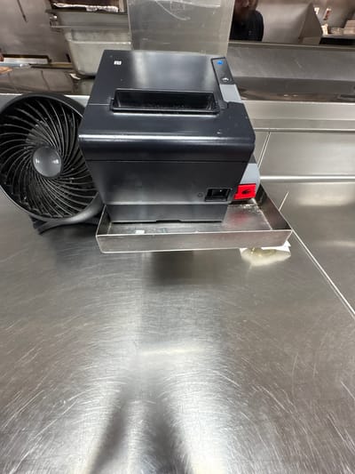 This media file captures a close-up view of a black electronic device, possibly a printer or scanner, mounted on a stainless steel surface. To the left of the device, a partially visible shelf holds what appear to be cleaning supplies. A person wearing a dark beanie is visible in the background, peeking over the shelf. Below the electronic device, a black fan with a circular grille is positioned on the stainless steel surface. The fan has a central circular emblem with text that is partially legible, appearing to read "Honeywell". The stainless steel surface exhibits numerous scratches, indicating wear and tear. The lighting suggests an indoor setting, possibly a commercial kitchen or utility area. There is a red lever with a white hand icon on the side of the electronic device, indicating a safety or operational feature. The scene appears to be set in Panama City Beach, USA. No specific activity is depicted, and the overall impression is one of a functional, everyday environment.
