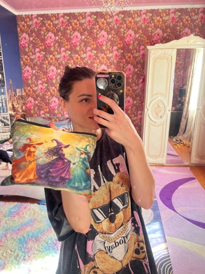 In a room with floral wallpaper and a pink ceiling, a woman is holding up a pouch featuring three witches in colorful dresses and pointed hats. She is holding her phone to take a picture of herself and the pouch in a mirror. In the background, a white ornate wardrobe and a purple rug are visible. The room appears to be a bedroom.