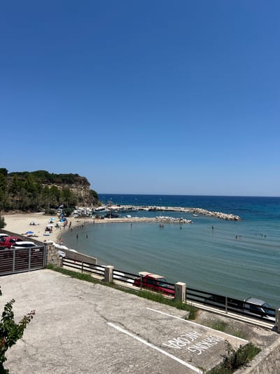 This media file captures a scenic view of a beachside parking area overlooking a bustling beach and a calm sea. The image is oriented vertically, suggesting it might have been taken with a phone held in portrait mode.

The primary subjects include people enjoying a day at the beach, with many visible in the shallow water, on the sand, and near some umbrellas. Various objects are present, such as parked cars, boats docked in the harbor, and protective barriers along the parking area and the waterfront. The scene is set in a coastal location, characterized by a sandy beach, a rocky breakwater extending into the sea, and a vegetated cliff face on the left side of the image. The setting appears to be a popular tourist destination.

The activity taking place is a typical summer day at the beach, with people swimming, sunbathing, and relaxing. The time of day is clearly daytime, indicated by the bright sunlight and clear blue sky, suggesting fair weather. The presence of a "PARKING ANCORA" sign, written in white on the concrete, provides a textual cue about the location, possibly indicating a designated parking area or a specific point of interest. The overall mood conveyed is one of leisure and enjoyment on a pleasant summer day.